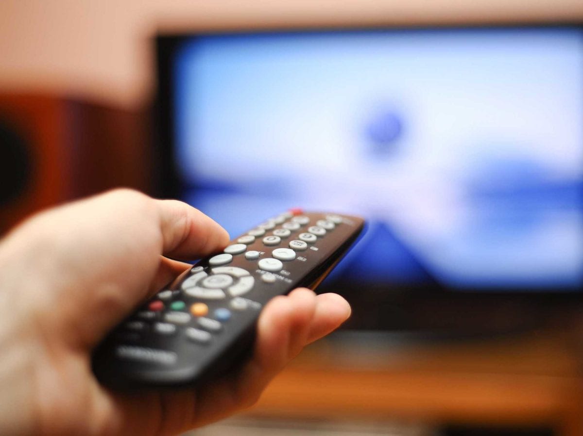 In the softly lit room, a hand grips the remote control as if wielding the fiery reins of a phoenix, guiding it toward the flickering television screen. The scene conjures an image of intricate flames dancing in harmony, mirroring the precise design of a well-crafted web interface where technology and comfort merge seamlessly.