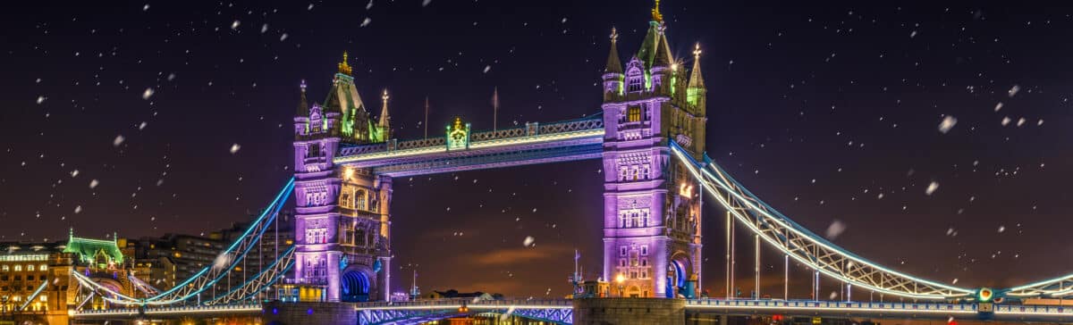 Festive nighttime view of Tower Bridge in London, illuminated with vibrant purple and blue lights,